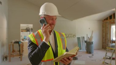 Bir inşaat yöneticisi, yoğun bir sitedeki planları gözden geçirirken telefonla etkili bir şekilde iletişim kurar ve yenilemelerde sorunsuz işlemleri sağlamak için profesyonellik ve çoklu görev becerileri gösterir