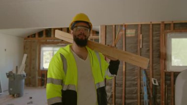 Güvenlik ceketi ve miğferi olan kendini işine adamış bir inşaat işçisi yenilenmiş bir iç mekanda kendinden emin bir şekilde kereste tutuyor ve takım çalışmasını ve inşaat sektörüne olan bağlılığını gösteriyor.