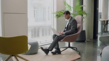 Son derece odaklanmış iyi giyimli iş adamı şık bir koltukta rahat bir şekilde oturuyor, dizüstü bilgisayarı üzerinde çalışmayı çağdaş bir ofis ortamında bitirmiş ve yerini terk ediyor.
