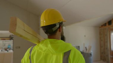Güvenlik kaskı ve yansıtıcı ceket giyen kendini işine adamış inşaat işçisi, yenilenmiş bir alanda kereste taşıyor ve bu da inşaat sürecine güçlü bir bağlılık ve proje başarısı gösteriyor.