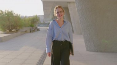 Gözlüklü profesyonel kadın, elinde dizüstü bilgisayarla modern şehir ortamında güvenle yürüyor, pozitiflik ve hırs yayarak kariyeri ve özel hayatı arasındaki dengeyi gösteriyor.