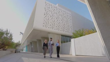 Çağdaş bir binanın dışında yürüyen ve fikirleri tartışan üç farklı iş adamı güneşli bir günde takım çalışmasını ve yeniliklerini destekliyor.
