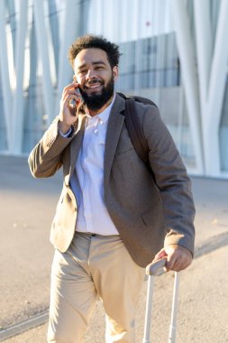 Şık giyinmiş neşeli bir gezgin şehrin sokaklarında şık bir bavulla rahatça yürüyor, telefonda modern mimarinin arka planına karşı canlı sohbetler ediyor.