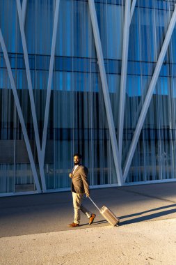 Kendine güvenen bir iş yolcusu hareketli bir şehirde, arkasında etkileyici modern mimari ile dolu şık bir bavulu manevra yaparak, profesyonelliği ve zarafeti somutlaştırır.