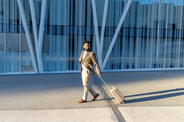 Çok şık ve kendine güvenen bir iş adamı, modern bir binanın önünde elinde bavulla kasıtlı olarak yürüyor. Bu da profesyonel seyahatin özünü gösteriyor.