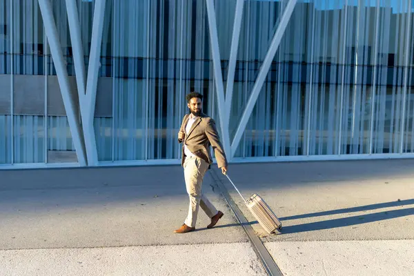 Çok şık ve kendine güvenen bir iş adamı, modern bir binanın önünde elinde bavulla kasıtlı olarak yürüyor. Bu da profesyonel seyahatin özünü gösteriyor.