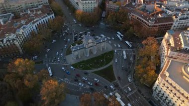 Madrid 'de ikonik Puerta de Alcal' ı gösteren çarpıcı hava aracı görüntüleri, gün batımında güzelce aydınlatılıyor, canlı sonbahar yeşillikleri ve araba trafiğiyle çevrili.