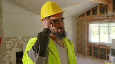 Sıkı bir şapka ve parlak bir güvenlik yeleği giyen kendini işine adamış bir inşaat işçisi, yoğun bir iş sahasında güven içinde el kol hareketleri yaparak, iş ortamını gözler önüne sererken önemli güvenlik önlemlerini vurguluyor.