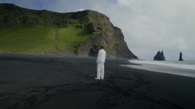 Beyazlar içindeki yalnız adam Reynisfjaras 'ın siyah kumları üzerinde duruyor. Etrafı çarpıcı kayalıklarla ve çarpan dalgalarla çevrili. İzlanda' yı somutlaştıran doğal güzellikler ve eşsiz manzaralar.