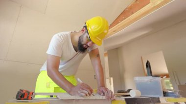 Korunmak için güvenlik kaskı takan kendini işine adamış ve odaklanmış bir inşaat işçisi titizlikle ahşap materyali ölçüyor.