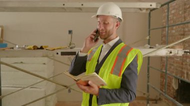 Sıkı bir şapka ve güvenlik yeleği giymiş kendini işine adamış bir profesyonel, yoğun bir inşaat sahasındaki notları inceleyerek, operasyonların sorunsuz geçmesi ve güvenlik ve verimliliğin korunması için görev başında bulunuyor.