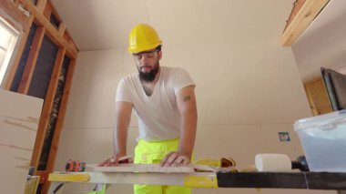Kendini adamış bir inşaat işçisi, önemli bir yenileme projesi için ahşap tahtaların hassas ölçümleri üzerinde yoğunlaşırken, aynı zamanda iyi aydınlatılmış bir çalışma alanı içinde gerekli güvenlik donanımını giyiyor.