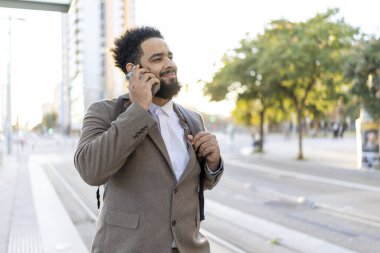 Keskin bir iş elbisesi giyen iyi giyimli profesyonel bir adam, insanlarla dolu hareketli bir şehir ortamında sıcak ve güneşli bir günün tadını çıkarırken canlı bir telefon görüşmesi yapıyor.
