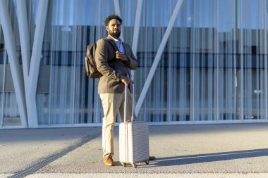 Profesyonel bir adam hem şık bir sırt çantası hem de sağlam bir bavul ile modern ve hareketli bir havaalanı terminalinin önünde duruyor. Bir sonraki yolculuğuna ya da macerasına hazır.