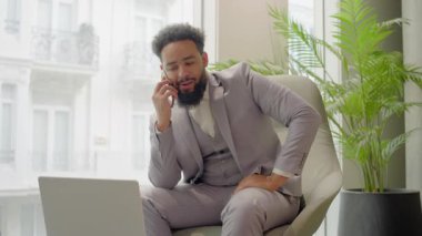 Yeşilliklerle dolu canlı ve iyi ışıklandırılmış bir çalışma alanında, takım elbiseli Afrikalı Amerikalı bir işadamı aynı anda bir telefon görüşmesiyle meşgul olurken aynı zamanda dizüstü bilgisayarı üzerinde çalışıyor.