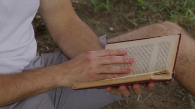 Şehir parkında okuyan ve ders çalışan tanınmamış genç öğrencilerin yakın görüntüsü. Beyaz erkek elleri bir kitap tutarken sayfaları çeviriyor. Yüksek kalite 4k görüntü