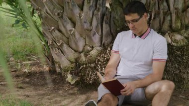 Gözlüklü, elinde roman tutan ve ağaca yaslanan beyaz bir adamın portresi. Şehir parkında okuyan ve okuyan genç bir öğrenci. Yüksek kalite 4k görüntü