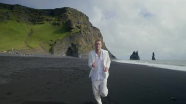 Beyaz atletizm kıyafetleri içindeki adam Reynisfjara sahillerinde enerjik bir şekilde koşuyor. Kayalıklar ve deniz yığınlarıyla çevrili, İzlanda 'nın çarpıcı manzaralarını ve doğal güzelliklerini ön plana çıkarıyor.