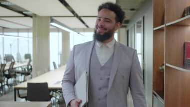 Afro-Amerikalı bir işadamı, şık giyinmiş, kendine güvenen, modern ve parlak bir ofis alanında yürüyor, güçlü bir profesyonellik ve hırs hissi yayıyor.