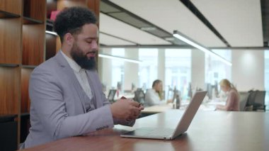 Hafif takım elbiseli bir Afrikalı Amerikalı laptopuna odaklanır. Çevresi çeşitli işlerde çalışan profesyonellerle çevrilidir. Dinamik bir atmosfere katkıda bulunur.