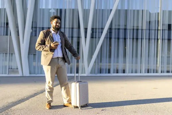 Kendine güvenen bir işadamı, iyi giyimli, havaalanı terminalinin önünde duruyor, elinde akıllı bir telefon ve şık bir bavul tutarken gülümsüyor. Bu resim seyahat ve iş bağlamlarına uyuyor