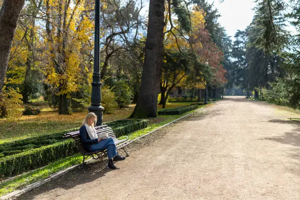 Huzurlu bir sahnede, bir kadın sakin bir parktaki bankta tek başına oturuyor, sonbahar yeşilliklerinin canlı renkleriyle çevrili, doğanın güzelliğine ve rahatlatıcı atmosfere dalmış.