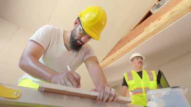 Yoğun bir inşaat sahasında, parlak başlıklı iki işçi karmaşık planları tartışırken, bir diğeri de devam eden projenin hassasiyetini güvence altına almak için sağlam bir tezgahtaki malzemeleri ölçüyor.