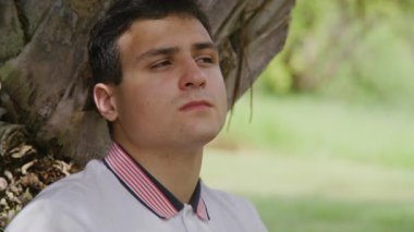 Zeki, genç bir adamın portresi güneşli bir sabahın ve yemyeşil parkın tadını çıkarıyor. Ağaca dayanmış düşünceli bir gencin yakın plan görüntüsü. Yüksek kalite 4k görüntü