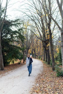 Bir kadın zarif bir şekilde, dingin ağaçlarla dolu sakin bir yolda yürüyor. Bu huzurlu sahnede ferahlatıcı sonbahar sükunetinin özünü mükemmel bir şekilde yakalıyor.