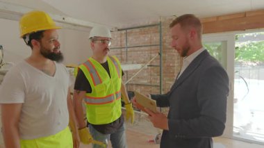 Bir işadamı inşaat işçilerinin çalışma alanındaki planlarını inceleyerek, takım çalışması ve iletişimi vurgulayan çağdaş bir yenileme projesinde işbirliğinin önemini vurguladı