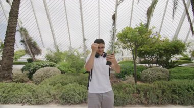 Yeşillik dolu egzotik şehir parkında yaz tatilinin tadını çıkaran bir turist. Kulaklıkları ve sırt çantasıyla dışarıda selfie çeken aktif bir genç. Yüksek kalite 4k görüntü