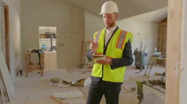 Koruyucu başlık ve parlak güvenlik yeleği giyen bir inşaat yöneticisi, her şeyin yolunda olduğundan emin olmak için evi baştan aşağı incelerken ayrıntılı notlar alır.