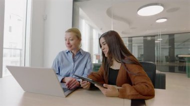 Dinamik bir ofiste, iki yetenekli kadın bir dizüstü bilgisayar ve tablet kullanarak işbirliği yaparak, modern işyerinde başarı ve yenilik için takım çalışması ve çeşitliliğin önemini vurguladılar.