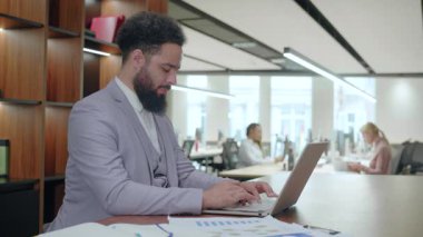 Başarılı bir Afrikalı Amerikalı işadamı modern bir dizüstü bilgisayarın başında şık bir iş yerinde oturuyor ve günümüz dinamik işgücünün üretkenliğini ve profesyonelliğini somutlaştırıyor.