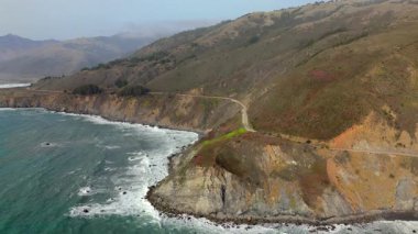 Bir hava görüntüsü, Kaliforniya kıyıları boyunca Big Sur 'un nefes kesen manzarasını yakalıyor. Otoyol 1' i gösteriyor. Bir araba, doğal güzelliği zengin, engebeli manzaralarda yol alırken.