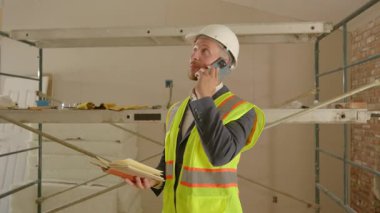 Güvenlik yeleği ve sert şapkalı bir inşaat yöneticisi elinde bir defter tutarken önemli bir telefon görüşmesi yapıyor. Devam eden bir inşaat projesini detaylara büyük özen göstererek dikkatle denetliyor.