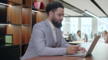 Canlı bir çalışma alanında, hafif takım elbiseli Afrikalı Amerikalı bir işadamı dizüstü bilgisayarında çalışırken, çeşitli meslektaşlar yaratıcı bir işbirliği içinde çalışarak dinamik bir çalışma ortamı sergiliyorlar.