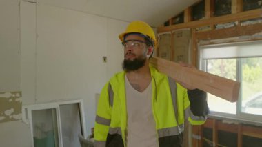 Uygun güvenlik teçhizatı giymiş bir inşaat işçisi ahşap bir kalas tutuyor. Tamamlanmamış bir iç mekâna dikkatle bakıyor, elimizdeki proje için bir sonraki adımları düşünüyor.