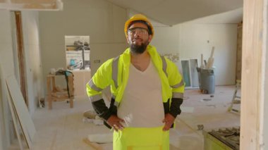 Kendine güvenen bir inşaat işçisi hareketli bir iş sahasında durur, gerekli güvenlik malzemelerini giyer ve dinamik çalışma ortamını ve üstesinden geldiği günlük zorlukları yansıtır.