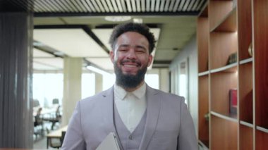 Canlı bir çalışma alanında, kendinden emin bir Afrikalı Amerikalı işadamı karizma ve profesyonellik yayar, dinamik bir çalışma ortamındaki yaklaşımını vurgulamak için içten bir gülümseme sergiler.