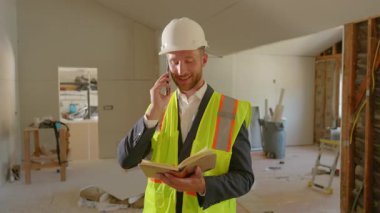 Sıkı bir şapka ve güvenlik yeleği giymiş bir inşaat yöneticisi önemli bir görevde, programa uyduğundan emin olmak için yenileme projesini denetliyor ve sıkı yönergeler ve protokollere bağlı kalıyor.