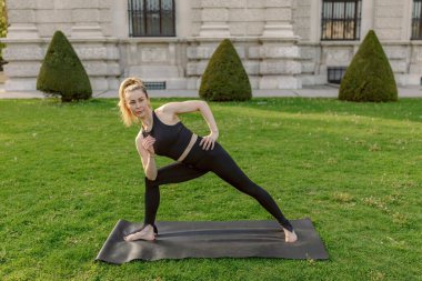 Güzel bir açık hava ortamında, midadlı bir kadın yoga yapar, esnekliğini ve gücünü gösterir spor yaparken ahenge değer veren fitness tutkunlarına ve doğa aşklarına seslenir.