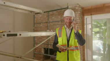 Sert şapka ve güvenlik yeleği giymiş bir inşaat uzmanı bir telefon görüşmesi yapıyor, bina planlarını gözden geçiriyor, arka planda canlı bir iç mekan ve iskelesi var, devam eden çalışmaları gösteriyor.