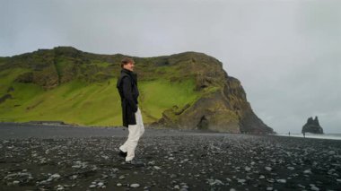 Tarz sahibi adam Reynisfjara plajı boyunca sakin bir şekilde geziniyor. Çarpıcı siyah kumları, yüksek uçurumları ve İzlanda manzarası boyunca nefes kesen okyanus manzaralarıyla ünlü ikonik bir yer.