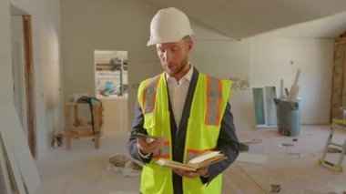 Yoğun bir inşaat sahasında, güvenlik yeleği ve başlık takan çalışkan bir inşaat yöneticisi, aynı zamanda her şeyin yolunda olduğundan emin olmak için telefonunu kullanarak defterine danışmanlık yapıyor.