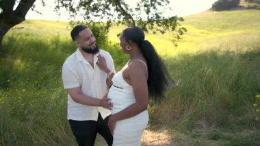 Neşeli, genç, hamile, Afro-Amerikan çift açık havanın tadını çıkarıyor. Derin sevgilerini ve pozitif ebeveynlik beklentilerini gösteren çarpıcı manzaranın tadını çıkarıyorlar.