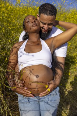 Afrika kökenli Amerikalı bir çift açık havada neşeli bir yaz gününün tadını çıkarırken, koca hamile karısına sarılıyor, doğanın güzelliği içinde mutluluklarını ve duygusal bağlarını kutluyor.