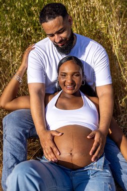 Mutlu ve neşeli hamile bir çift, dışarıda güzel bir yaz gününün tadını çıkarırken, koca, tatlı bir kutlama anında karısına şefkatle sarılıyor ve onu nazikçe öpüyor.