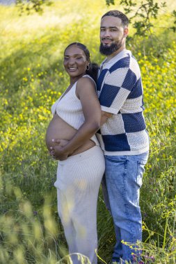 Sevgi dolu hamile bir çift açık havada güneşli bir yaz gününün tadını çıkarırken, koca, karısına nazikçe sarılıyor ve yemyeşil ve canlı çiçeklerle dolu huzurlu bir ortamda bebek çarpması yaşıyor.