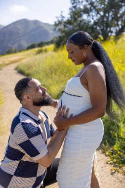 Sevecen bir Afrikalı Amerikalı çift, sıcak yaz mevsiminde dışarıda olmanın tadını çıkarıyor. Hamileliğin güzelliğini ve doğanın güzelliklerini birlikte kucaklıyorlar.